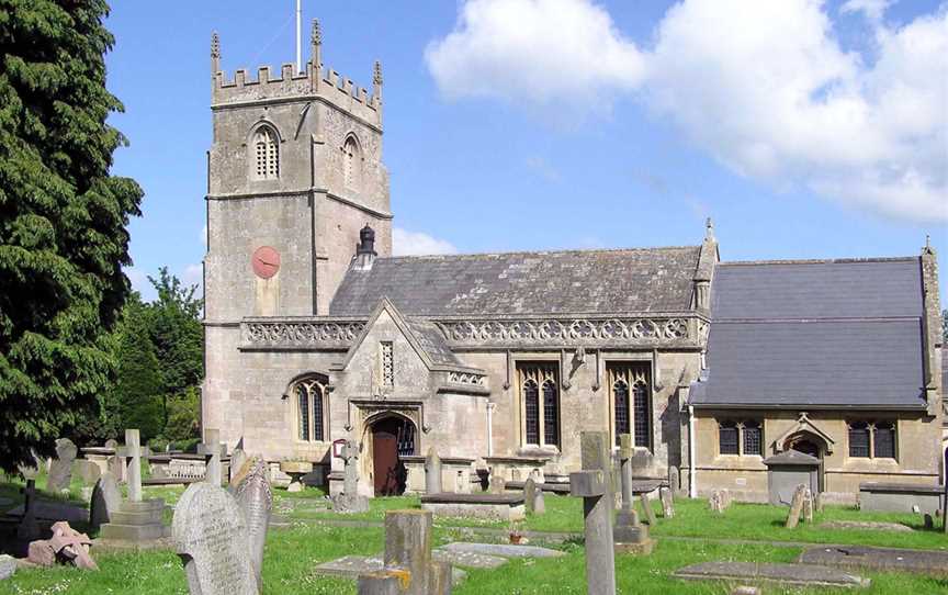 Yellow stone building, with porch with triangular roof in front. Short square tower with battlements topped by flag and flag pole. Gray gravestones in the foreground.