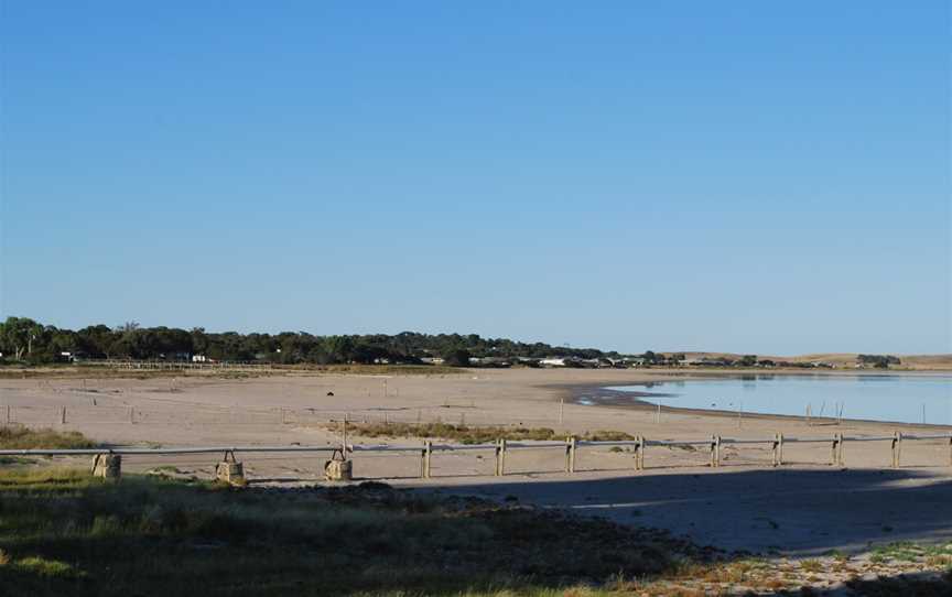 The picture shows Lake Albert from its shore. In the foreground is a sandy shore and a pipeline leading towards the lake. The lake is in the right of the photo. In the background there are trees on the left and brown hills on the right.