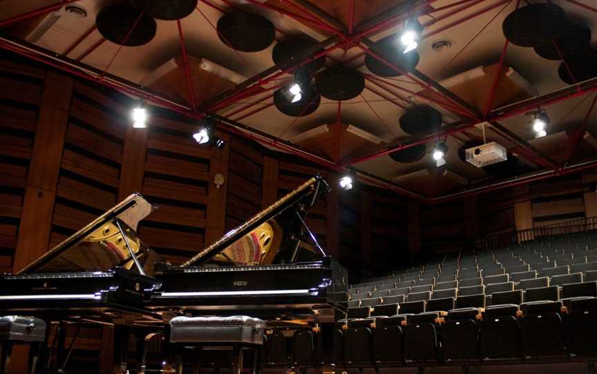 Two shiny black pianos with their lids open are side by side, and in the background is the ranked seating of a performance space