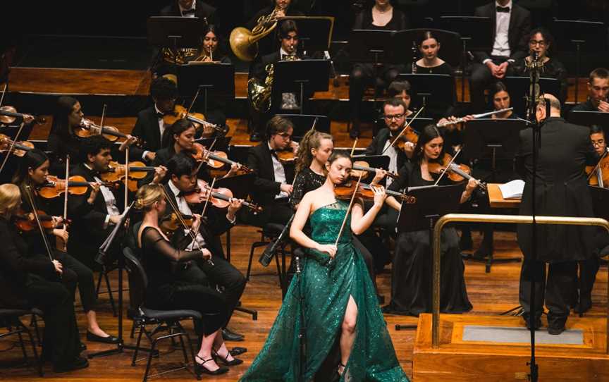 Woman in a sparkly green dress is playing her violin as a soloist in front of the orchestra.