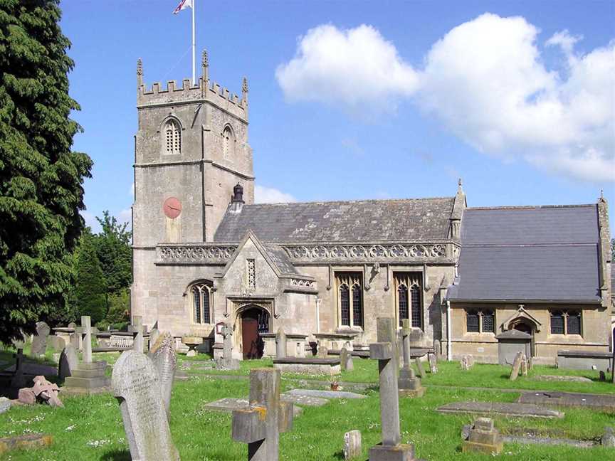 Yellow stone building, with porch with triangular roof in front. Short square tower with battlements topped by flag and flag pole. Gray gravestones in the foreground.