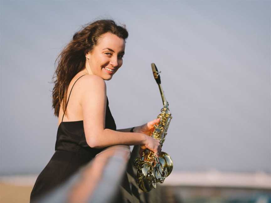 Emma McPhilamy is holding her shiny saxophone and looking to the side at the camera.