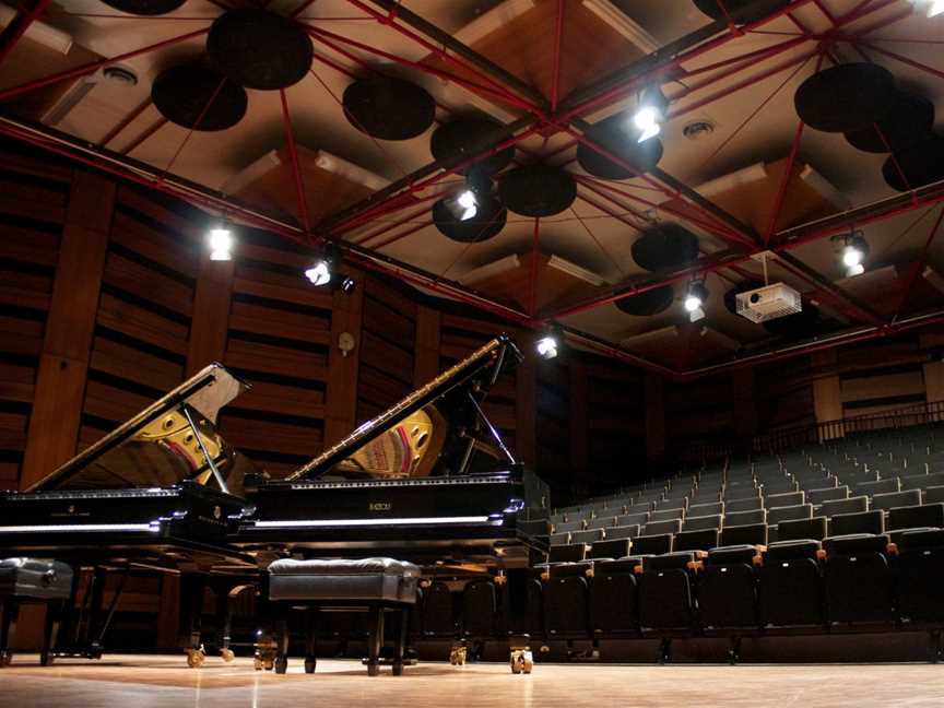 Two shiny black pianos have their lids opened and are standing side by side, in front of the ranked seating in a performance space.