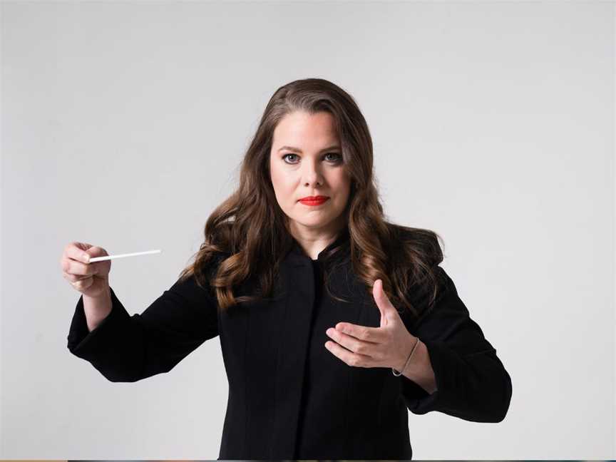 Conductor dressed in all black with red lipstick facing the camera with her baton mid-swing