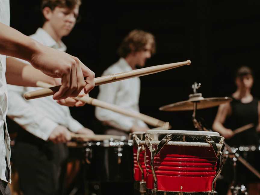Foreground shows a pair of hand holding wooden drumsticks which are hitting the head of a pair of bright red drums.