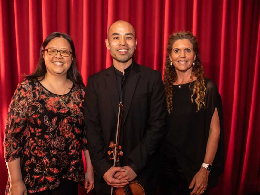 Three people stand smiling at the camera, in front of a red curtain. The man in the middle is holding his violin.